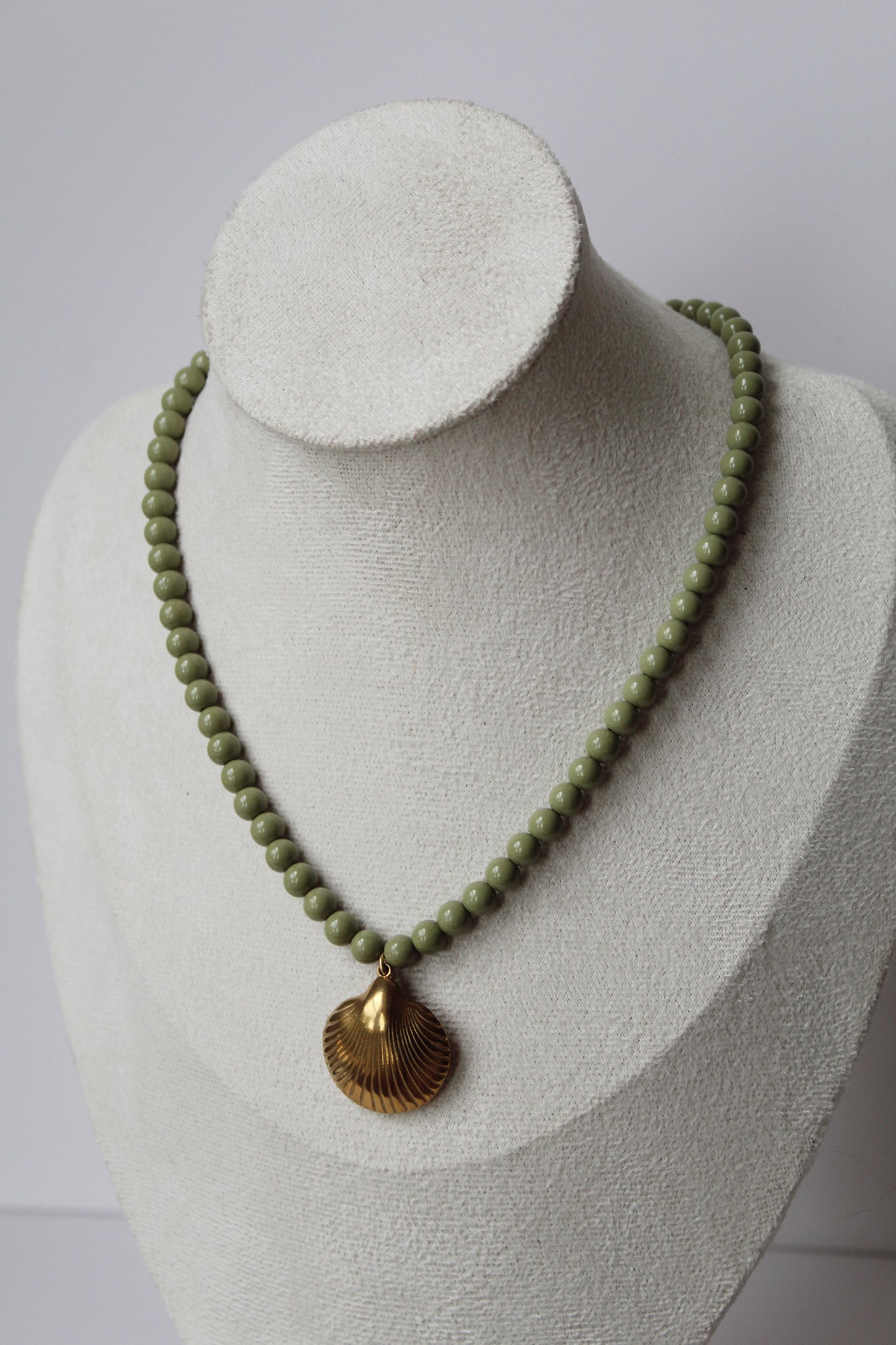 Sage green beaded necklace with a gold shell pendant displayed on a jewelry bust.
